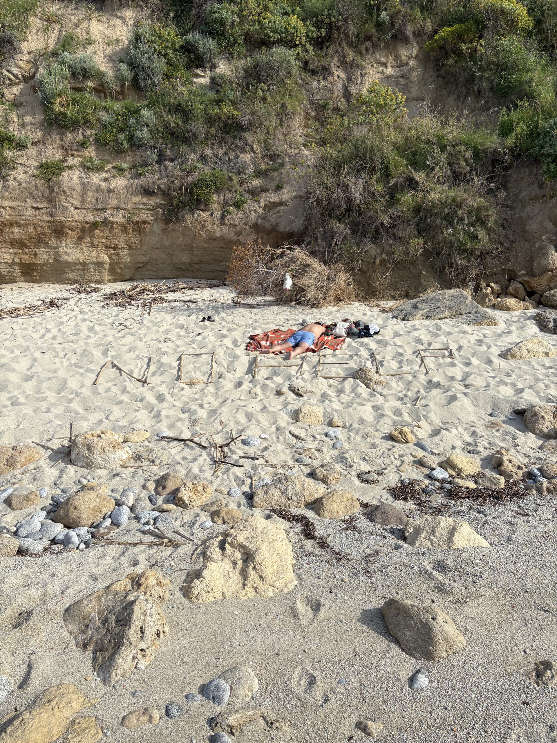 Piotr Jawornik leżący na dzikiej plaży z napisem NO HELP ułożonym z patyków — Kalabria — fot. © Piotr Jawornik